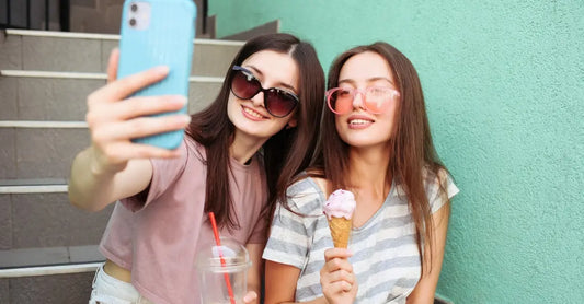 Two young women from Gen Z are taking a selfie while sitting on concrete stairs in front of a teal wall holding a coffee and an ice cream cone