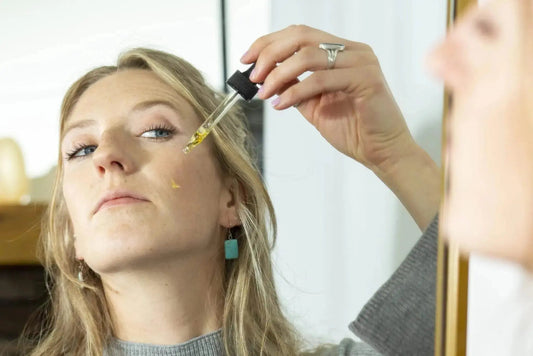A photo of a young woman looking in the mirror applying Simple Body Pomegranate Oil to her cheek with beautiful skin.