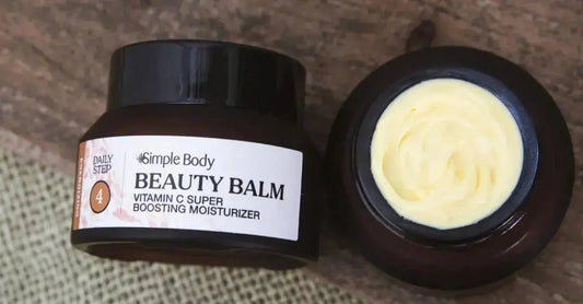 A jar of Simple Body Beauty Balm, Vitamin C Super Boosting Moisturizer. One jar is laying on it's side showing the label and the other jar has it's lid removed showing the creamy yellow texture of the Beauty Balm.