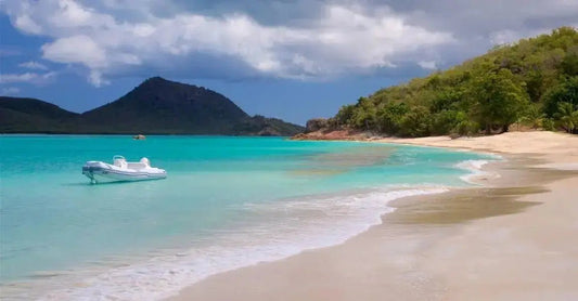 A photo of the beautiful blue teal waters of Antigua in the Carribean with a lone boat, sandy beach and green trees with mountains in the background.