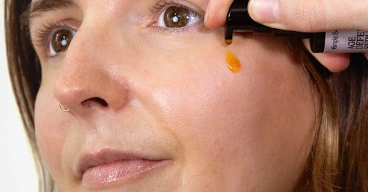 A close up image of Simple Body Age Defense Serum being applied to the cheek of a young woman with brown eyes. 