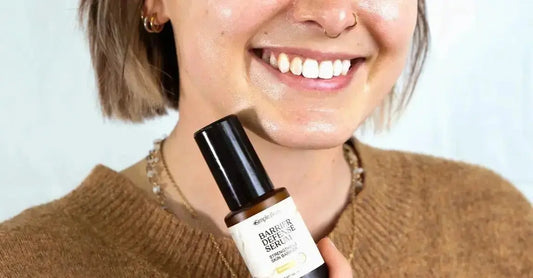 A close up photo of a young woman from her nose down, smiling, holding up a Simple Body Barrier Defense Serum with a rusty brown sweater.