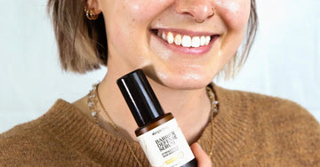 A close up photo of a young woman from her nose down, smiling, holding up a Simple Body Barrier Defense Serum with a rusty brown sweater.
