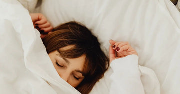 A young woman sleeping in a bed with white sheets and a white comforter with half her face buried under the covers.