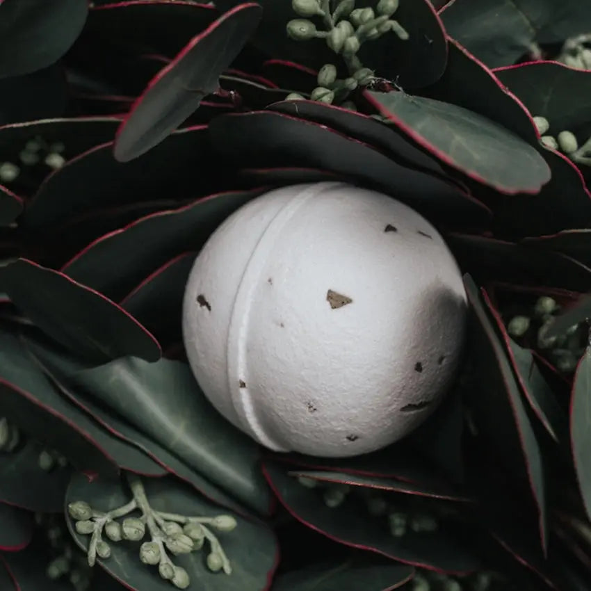 A photo of a white natural bath bomb with herbs in it on a leavy, dark green background.