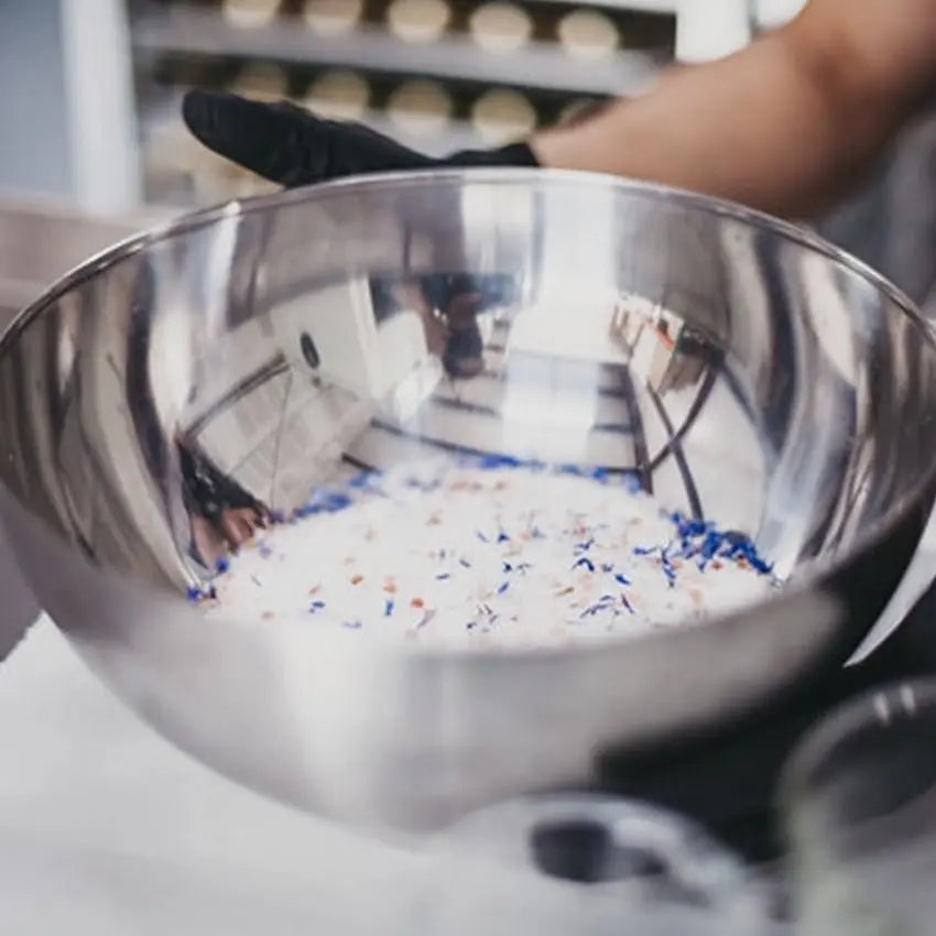A photo of gloved hands holding a silver bowl with natural bath salt in it and fresh purple and yellow herbs and a blurred background.