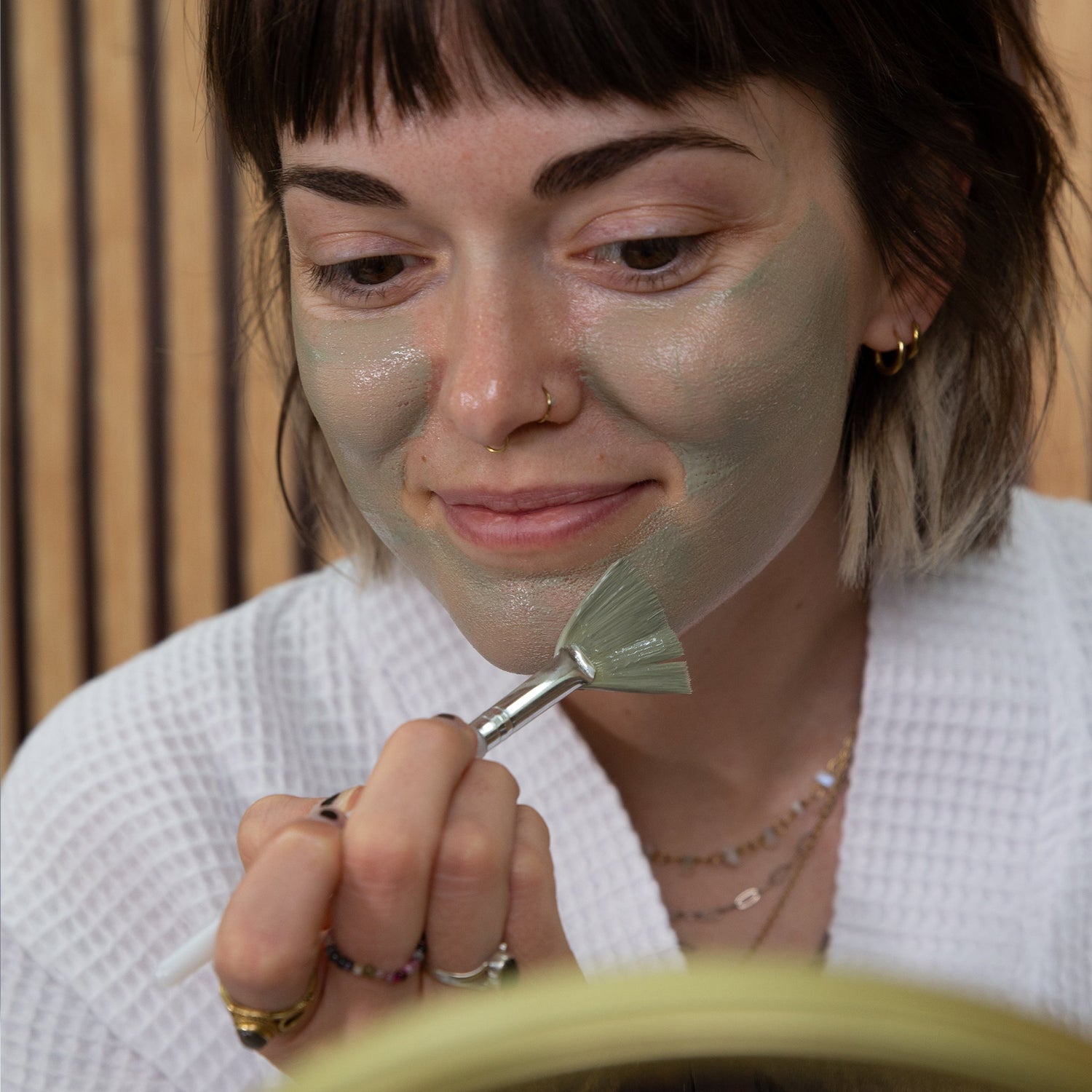 A photo of a young woman applying a green clay mask with a fan brush and wearing a white robe.