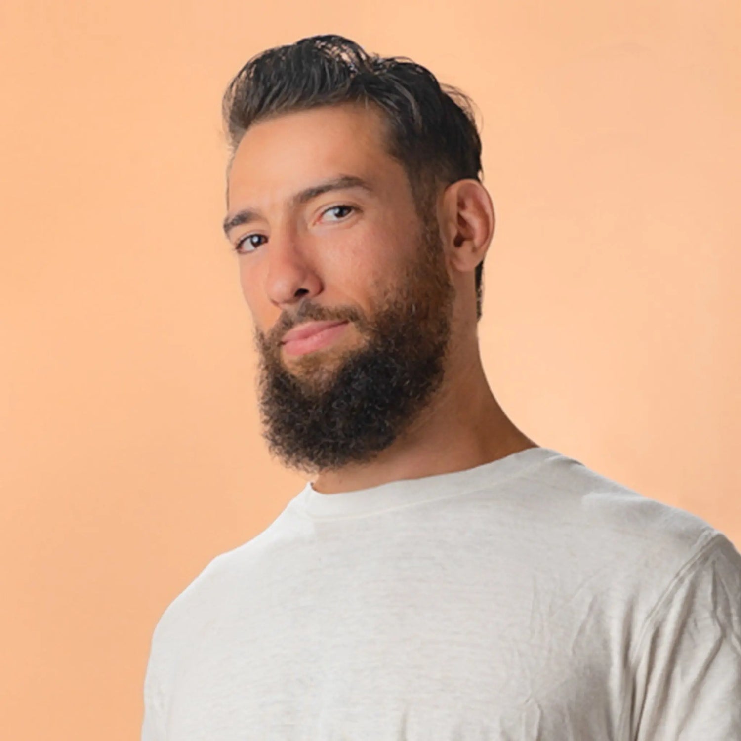 A handsome young man with a beard and dark brown eyes wearing a white tee shirt looking directly at you with a coy smile.