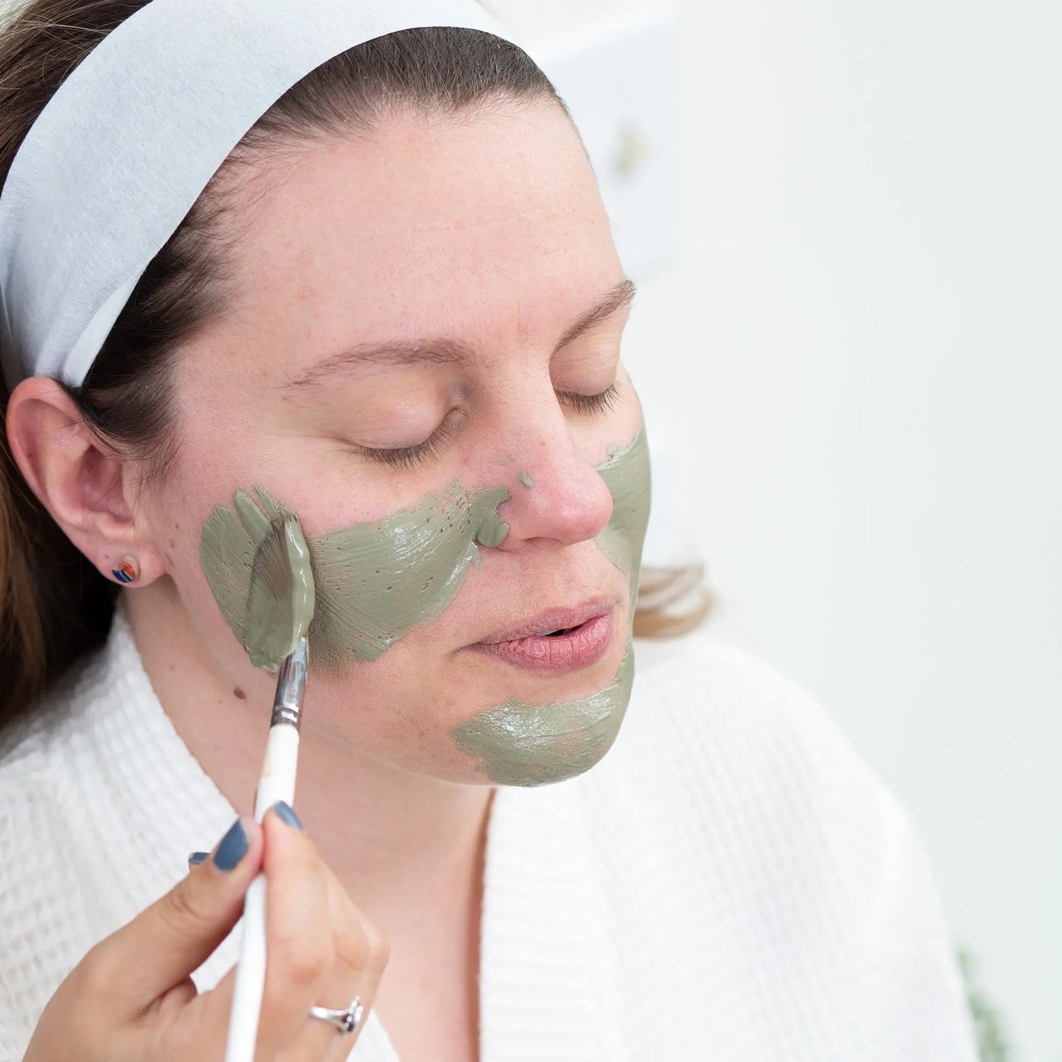 A photo of a woman with her eyes closed applying a Simple Body Green Clay Mask with a white headband and wearing a white waffle robe. 