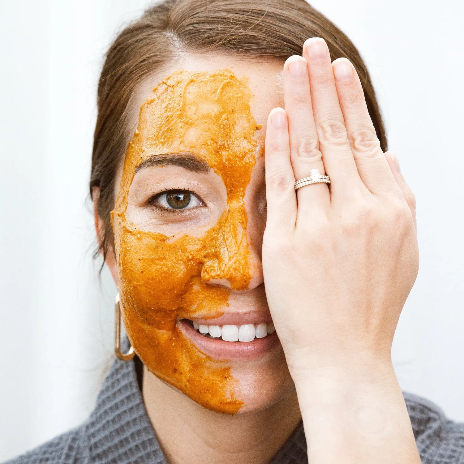 A photo of a beautiful young woman covering one side of her face and wearing a pumpkin enzyme mask on the other.k