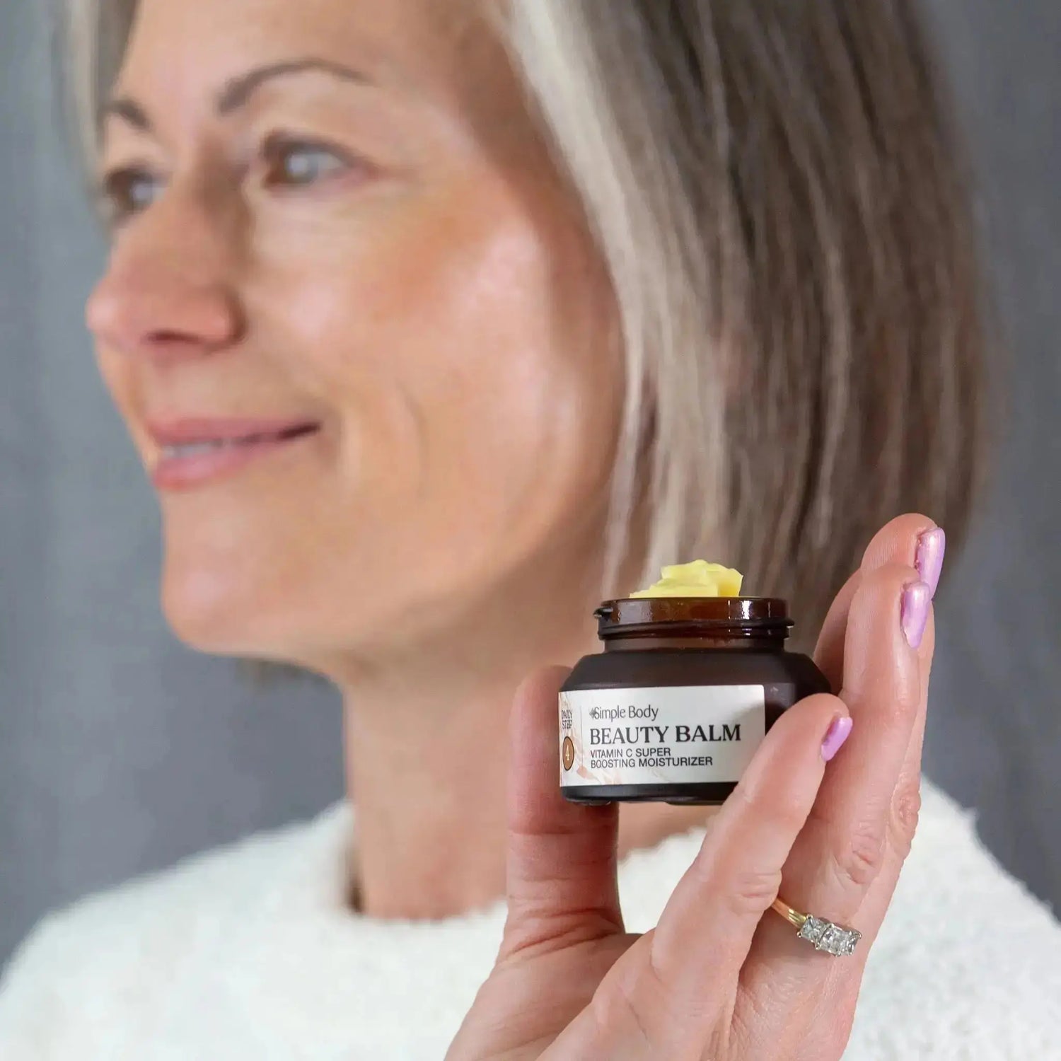 Michelle holds a jar of Simple Body Beauty Balm which shows a rich, yellow Vitamin C moisturizer in the jar. Michelles skin looks deeply moisturized and glowing. 