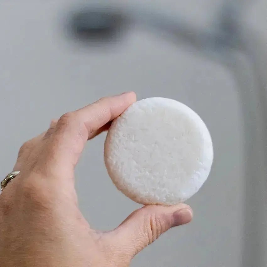A closeup image of a hand holding a white, natural shampoo bar that is slightly textured with a blurred background.
