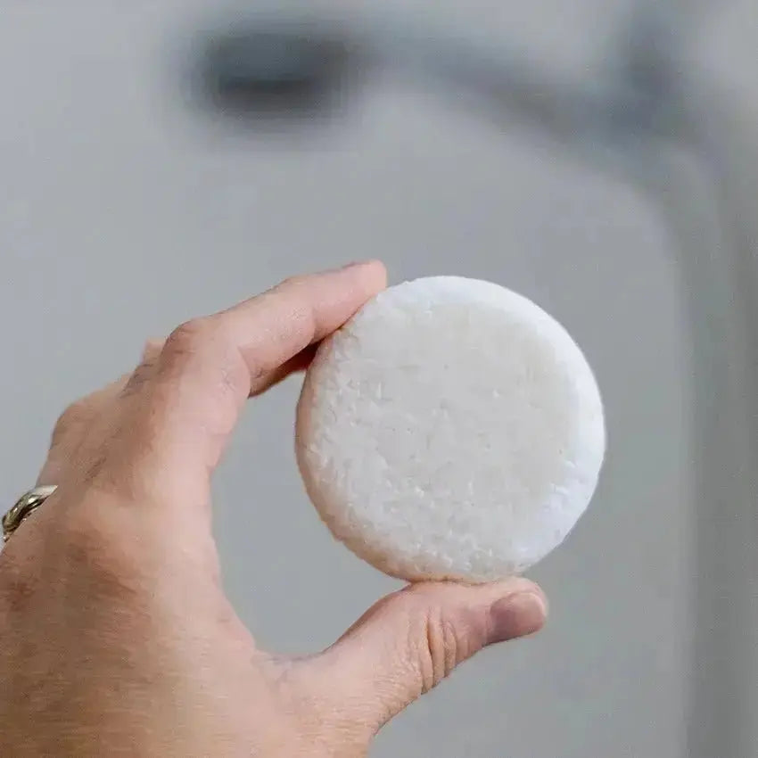 A closeup image of a hand holding a white, natural shampoo bar that is slightly textured with a blurred background.