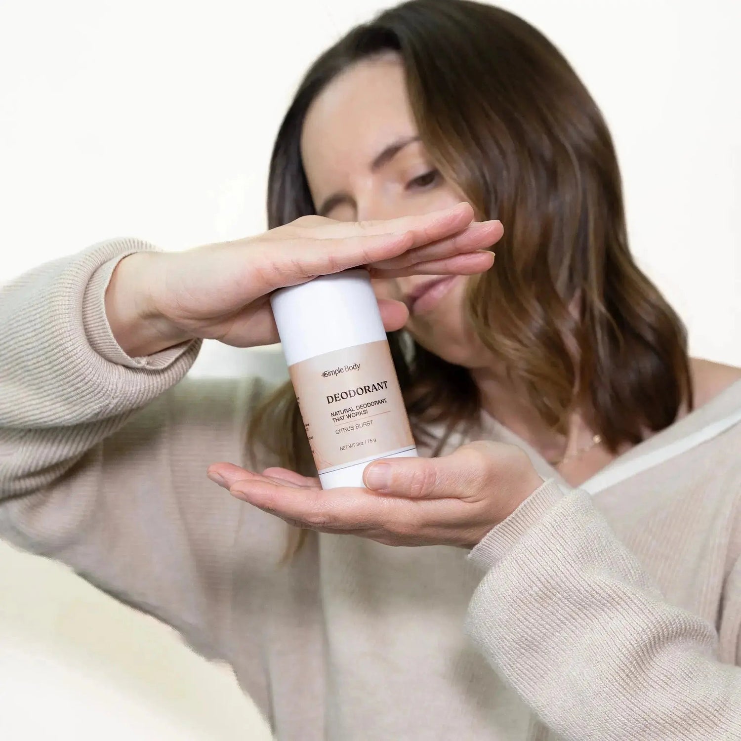 Ashley holds a stick of Simple Body Natural Citrus Burst Deodorant, part of the All-Natural Deodorant Collection. She gazes down at the deodorant as if reading the natural ingredients. She's wearing a beige lounge set.