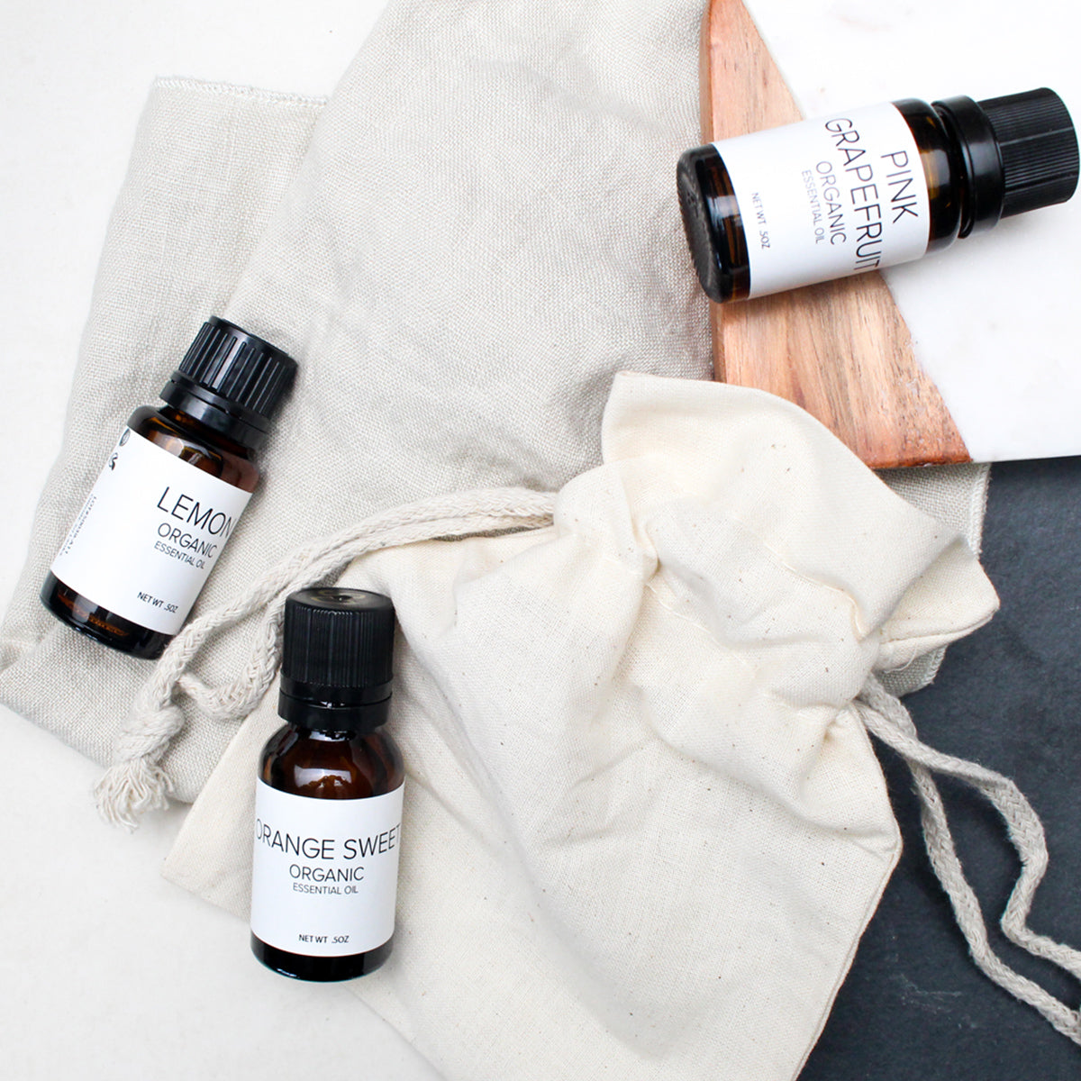 A photo of three amber colored bottles of essential oils with white labels including Lemongrass, Orange Sweet, and Pink Grapefruit laying on organic cotton bags and a wood and marble tile.