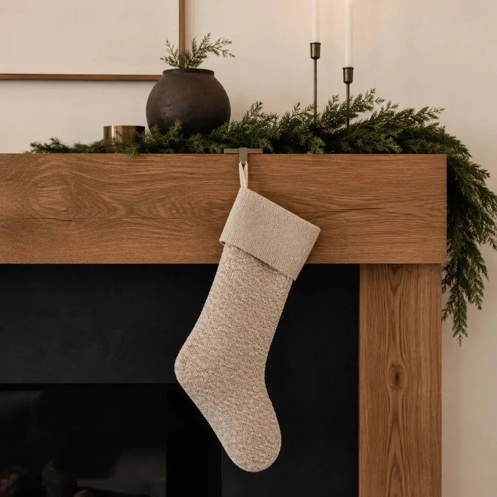 A minimal beige holiday stocking hangs from a beautiful wooden mantel with boughs of greenery, a brown vase and two candlesticks
