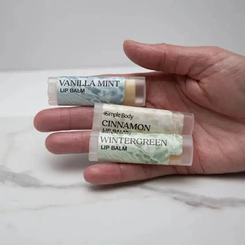A closeup photo of a female hand holding a Simple Body Lip Balm collection including Vanilla Mint, Cinnamon, and Wintergreen Lip Balms on a white marble background.