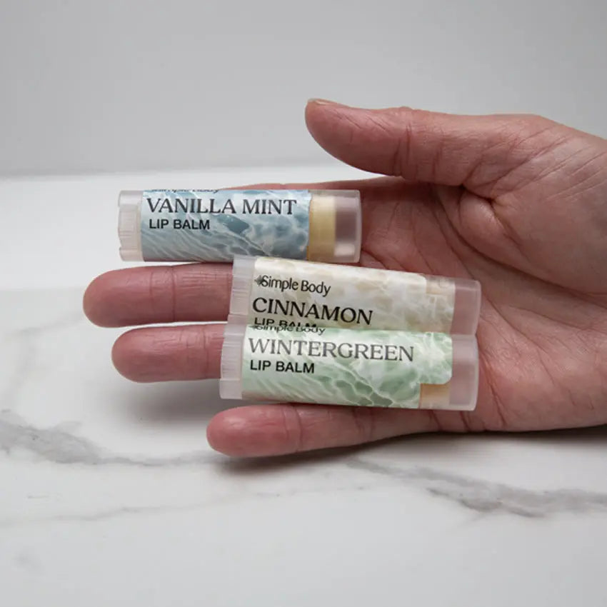 A closeup photo of a female hand holding a Simple Body Lip Balm collection including Vanilla Mint, Cinnamon, and Wintergreen Lip Balms on a white marble background.