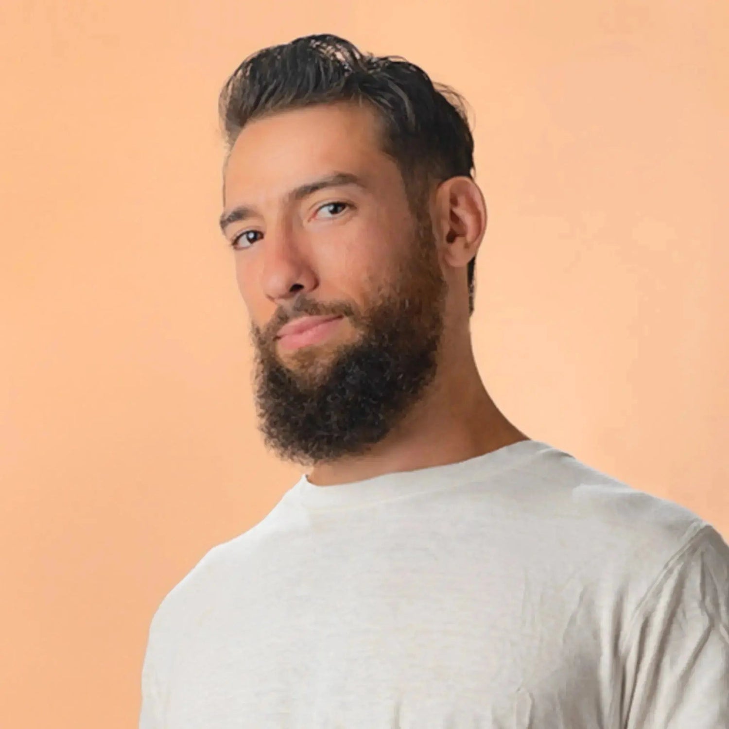 A handsome young man with a beard and dark brown eyes wearing a white tee shirt looking directly at you with a coy smile.