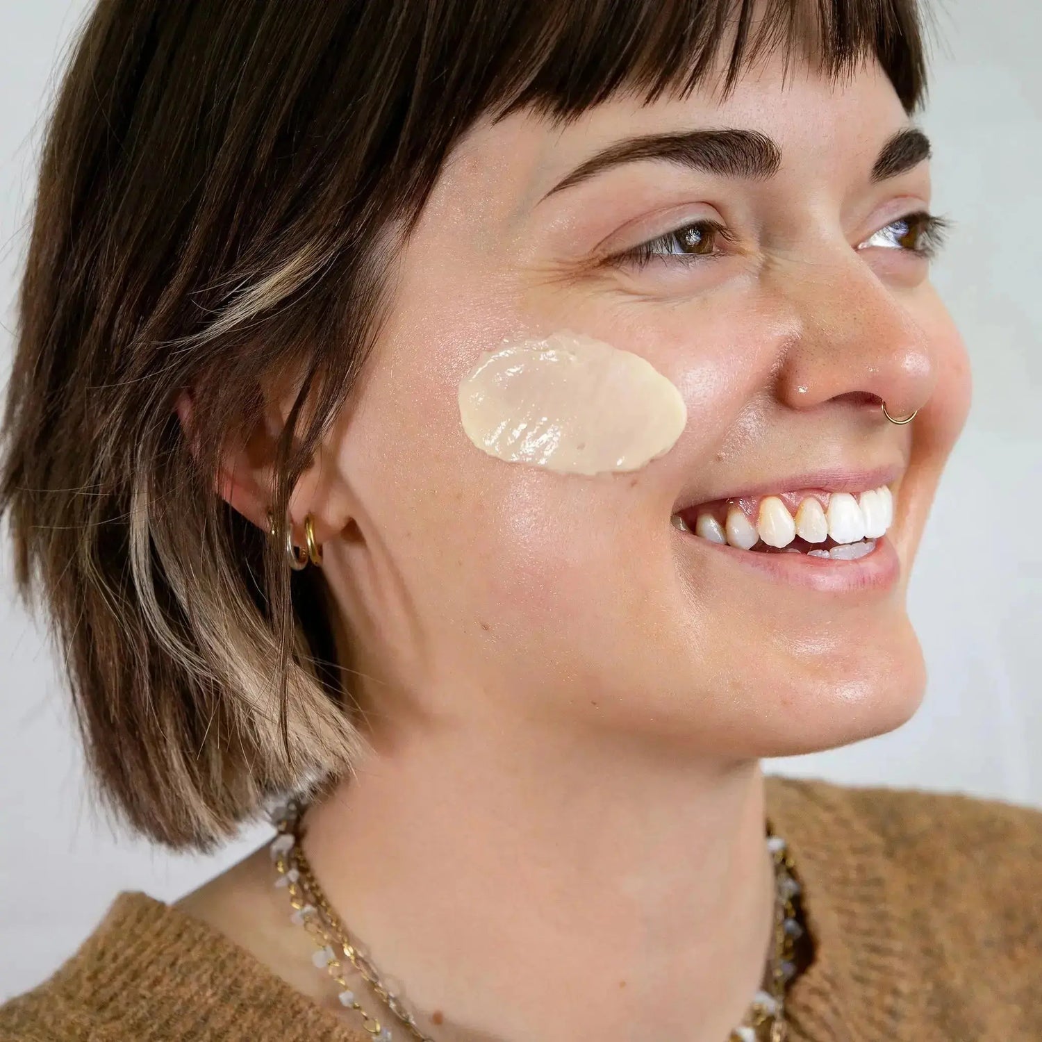Corrin smiles as she shows off a swatch of Simple Body Face Cream from the Moisturizers Collection on her cheek. She's very happy and wearing a tan sweater with cute jewelry.