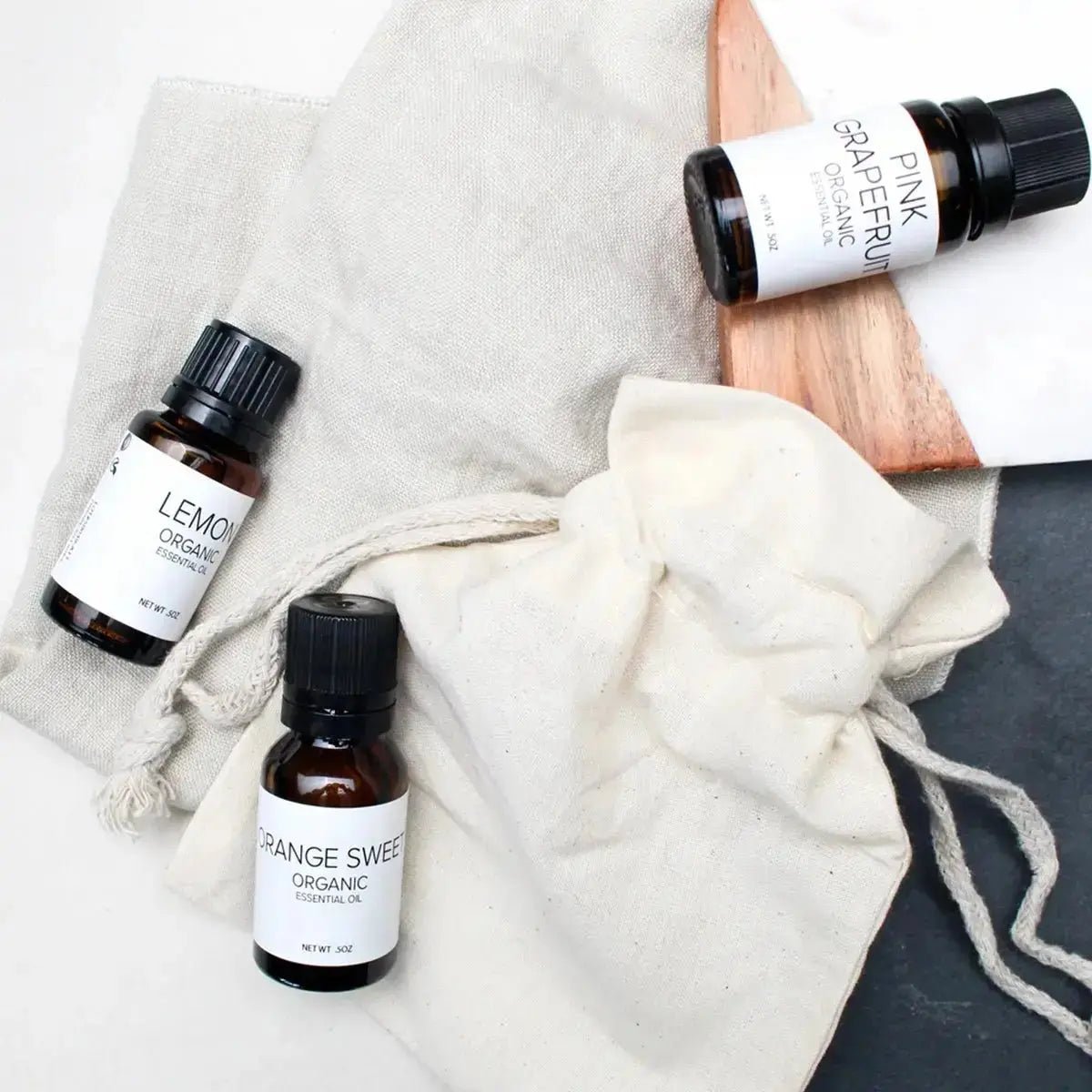 A photo of three amber colored bottles of essential oils with white labels including Lemongrass, Orange Sweet, and Pink Grapefruit laying on organic cotton bags and a wood and marble tile.