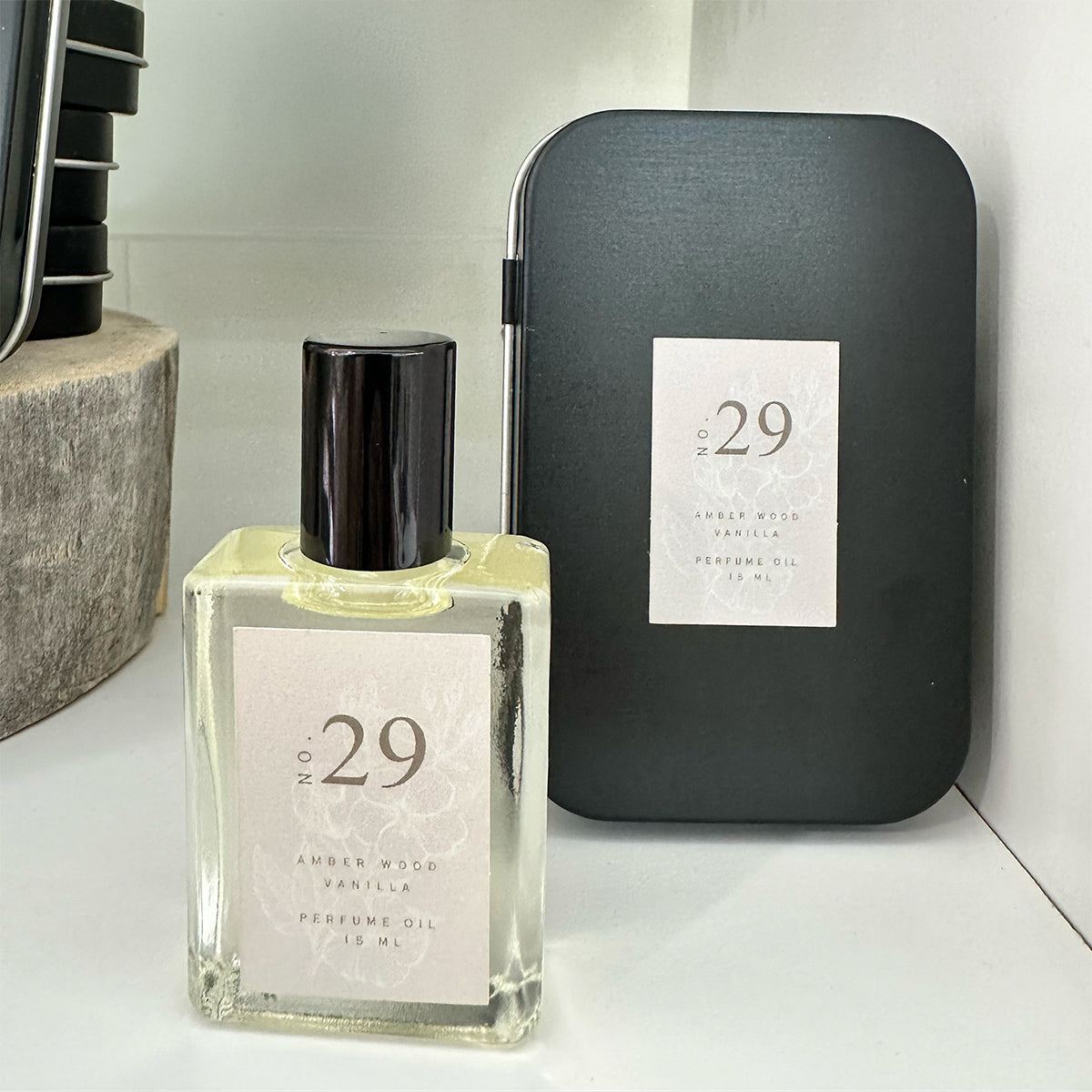 A photo of a no. 29 natural essential oil perfume roller in a glass bottle with a black top and a black tin behind it on a white background.