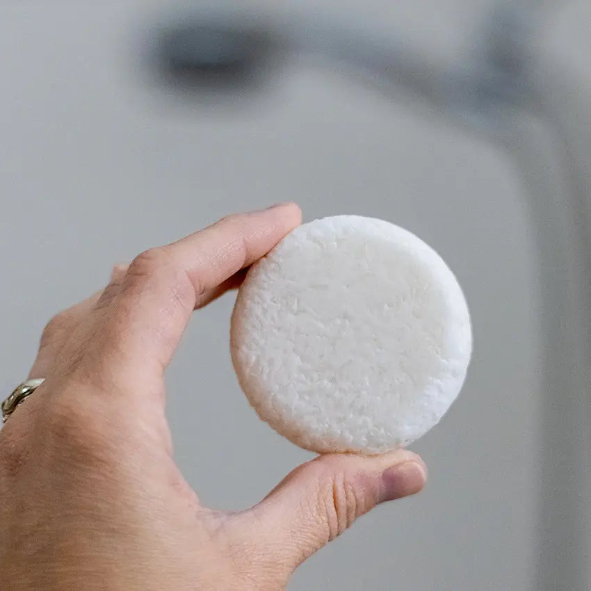 A closeup image of a hand holding a white, natural shampoo bar that is slightly textured with a blurred background.