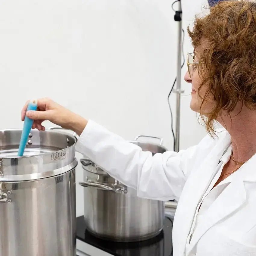 A photo of Simple Body Founder, Jewels Burdick in a white lab coat stirring handmade, natural skincare in a stainless steel pot.
