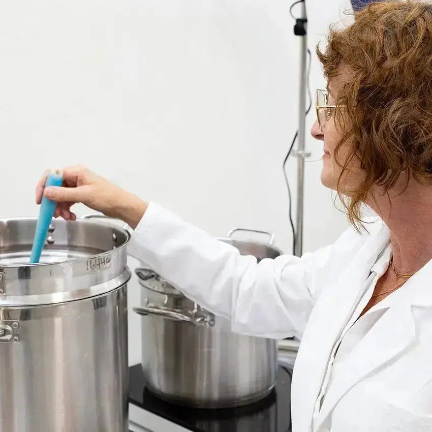 A photo of Simple Body Founder, Jewels Burdick in a white lab coat stirring handmade, natural skincare in a stainless steel pot.