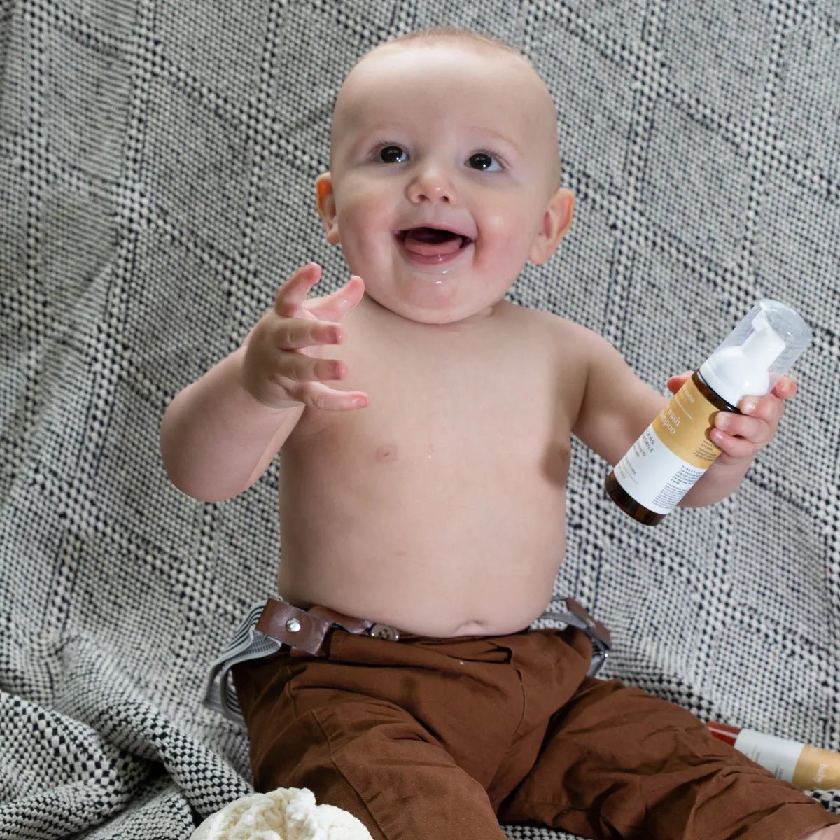 An adorable baby boy looks up and smiles holding a Simple Mama Baby Wash with a patterned background.