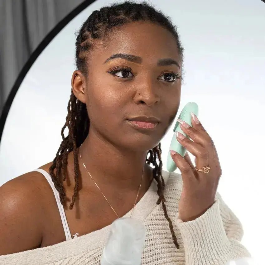 A photo of a young, black woman holding a Simple Body Facial Cleansing Brush with a white off the shoulder sweater on a white background.