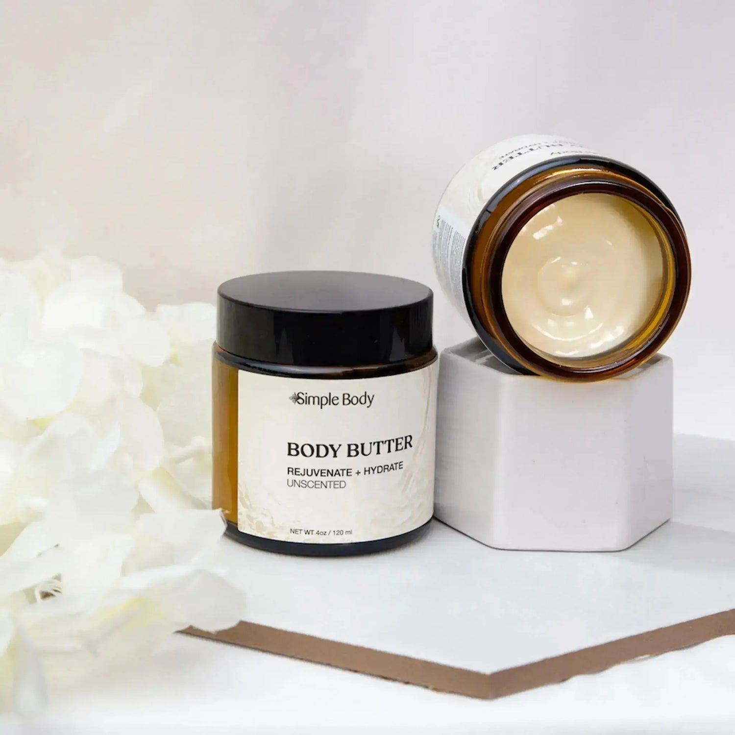A Simple Body Unscented Body Butter is propped on its side showing the creamy texture of the Body Butter and along side it is a freestanding Unscented Body Butter
