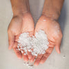 Two hands holding a small amount of Bloom Bath Co Eucalyptus and Lavender white bath salt on a neutral background