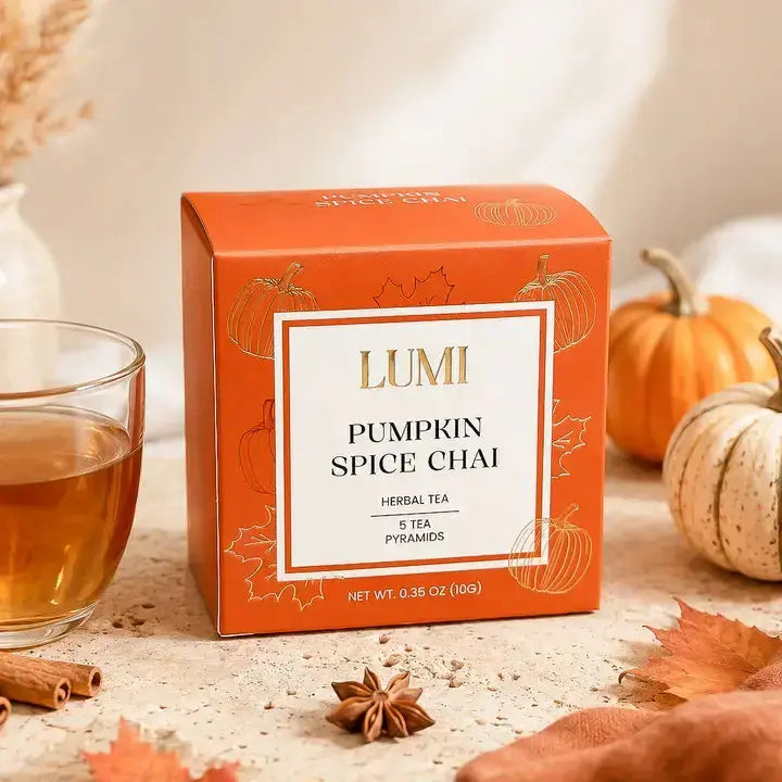 A box of Lumi Pumpkin Spice Chai herbal tea sits on a stone surface with a glass of tea, autumn leaves, cinnamon, star anise, and small pumpkins around it.
