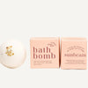 Two pink bath bomb boxes and a white round bath bomb with gold leaf flakes, arranged on a warm cream background. The left box reads 'bath bomb' and the right box reads 'sunbeam'; both say 'essentially ginger' and 'lightly'.