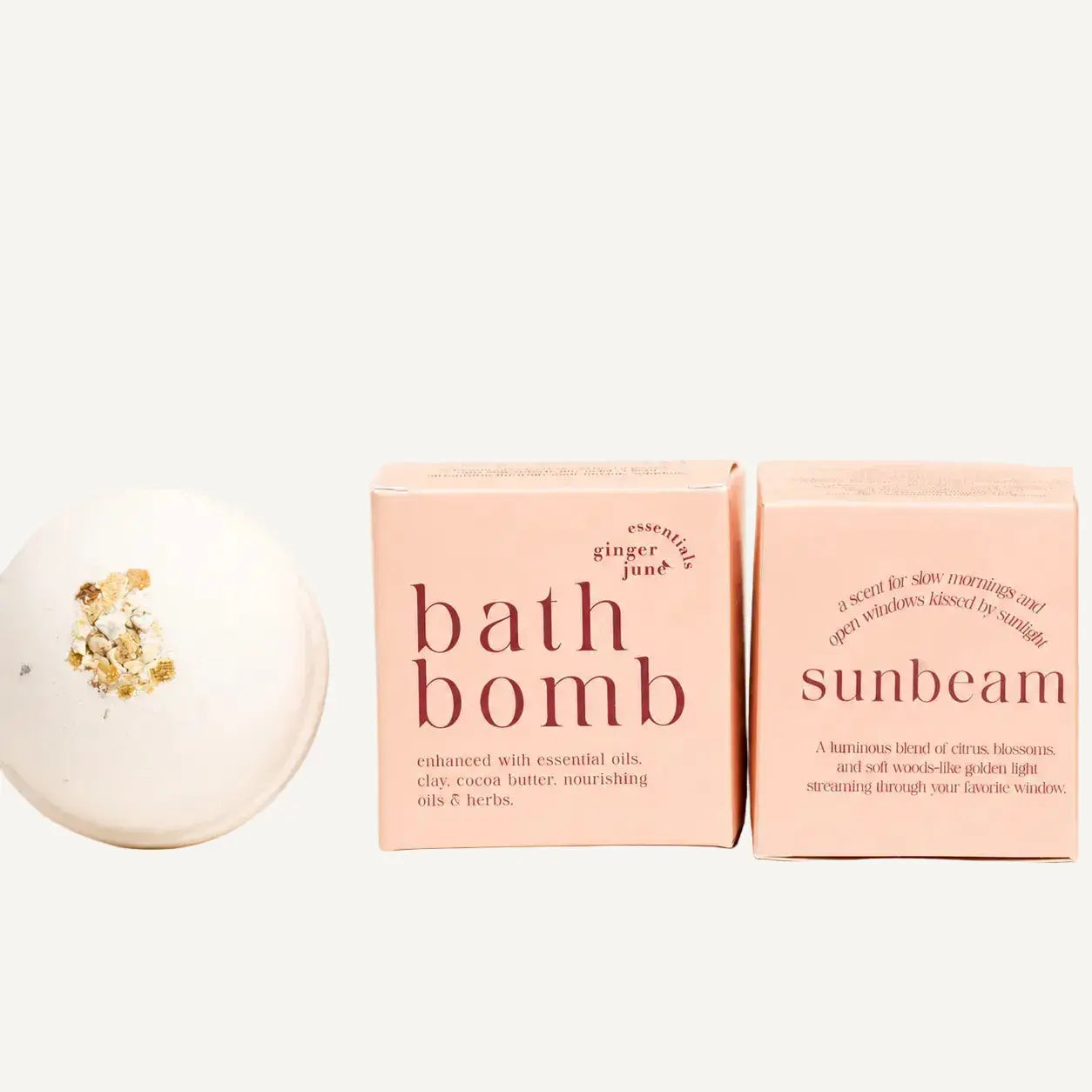 Two pink bath bomb boxes and a white round bath bomb with gold leaf flakes, arranged on a warm cream background. The left box reads 'bath bomb' and the right box reads 'sunbeam'; both say 'essentially ginger' and 'lightly'.