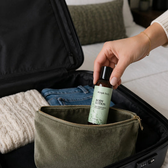 An olive green zippered cosmetic pouch sits inside an open black suitcase, with a hand placing a travel-sized Simple Body Rosemary Mint Body Lotion bottle into the pouch. Folded jeans and a knit sweater are visible in the suitcase, and a bed and pillow appear in the softly blurred background.