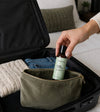 An olive green zippered cosmetic pouch sits inside an open black suitcase, with a hand placing a travel-sized Simple Body Rosemary Mint Body Lotion bottle into the pouch. Folded jeans and a knit sweater are visible in the suitcase, and a bed and pillow appear in the softly blurred background.