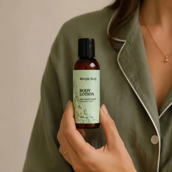 A person in an olive green pajama shirt holds a bottle of Simple Body Travel-sized Rosemary Mint Body Lotion. The lotion bottle is amber brown with a black cap and a light green label.