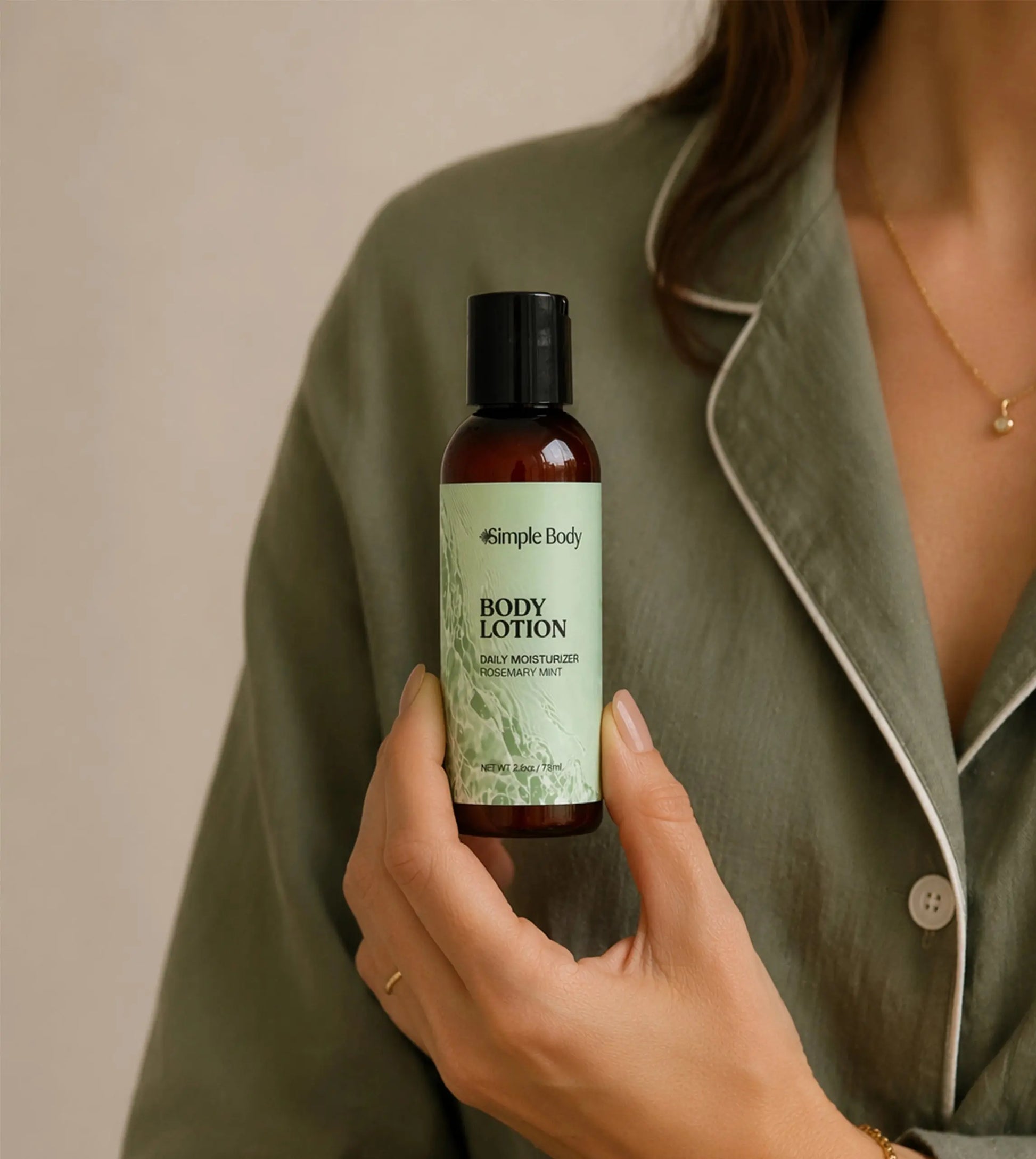 A person in an olive green pajama shirt holds a bottle of Simple Body Travel-sized Rosemary Mint Body Lotion. The lotion bottle is amber brown with a black cap and a light green label.