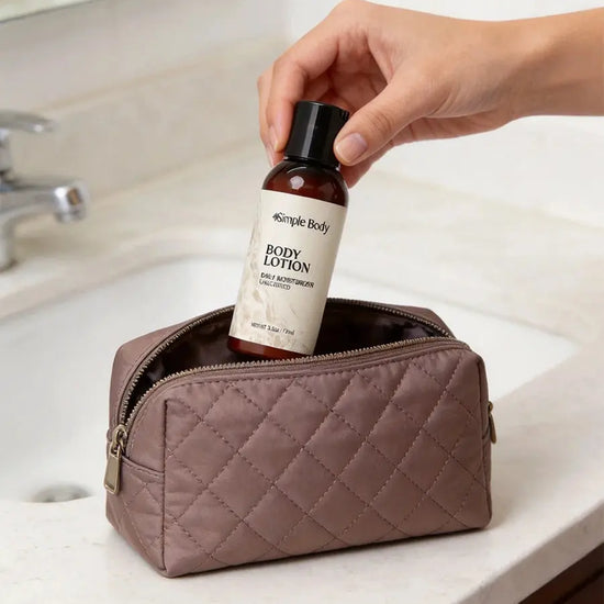 A woman's hand holding a bottle of Simple Body Unscented Body Lotion over a quilted tan toiletry bag on a bathroom counter.