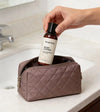 A woman's hand holding a bottle of Simple Body Unscented Body Lotion over a quilted tan toiletry bag on a bathroom counter.