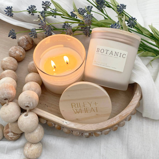 Two candles on a wooden tray with 'Botanic' and 'Riley + Wheat' branding, surrounded by lavender and wooden beads.