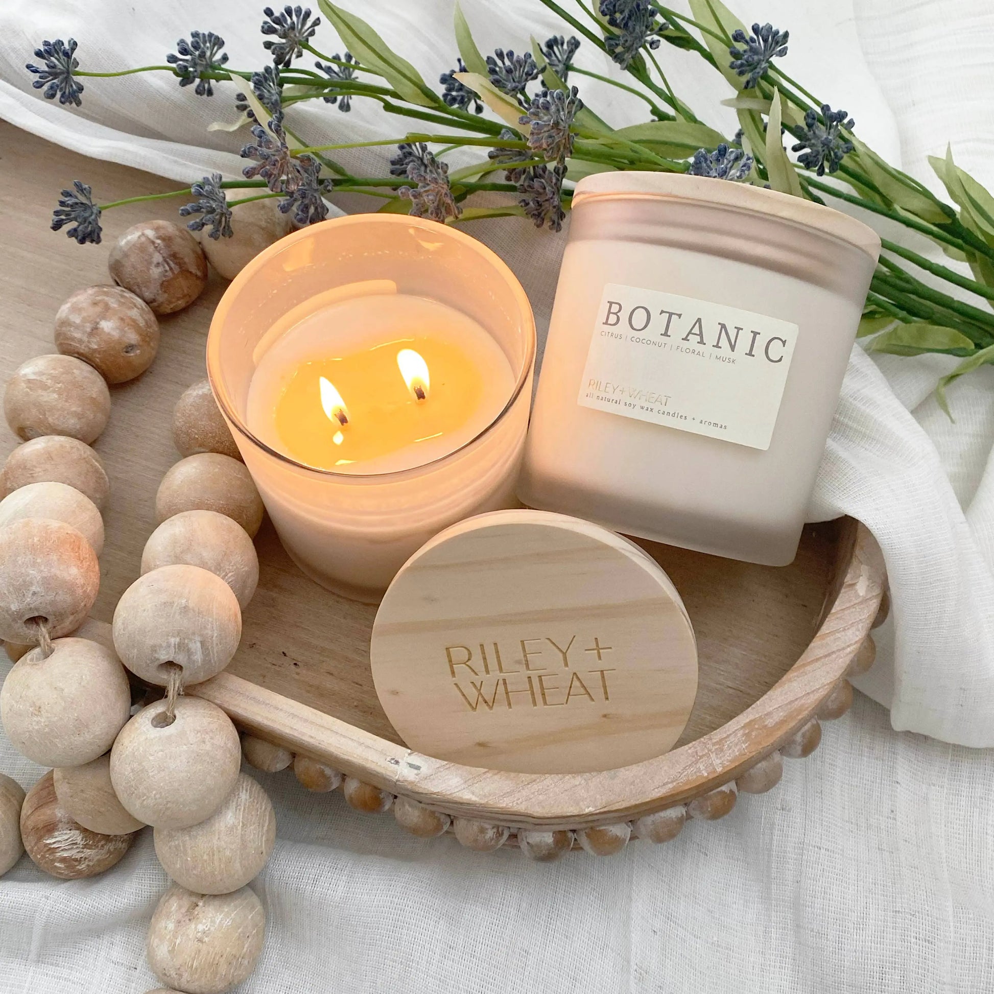 Two candles on a wooden tray with 'Botanic' and 'Riley + Wheat' branding, surrounded by lavender and wooden beads.