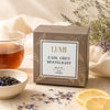 A box of LUMI Earl Grey Moonlight black tea sits on a light stone surface with a glass of brewed tea, a small bowl of loose tea leaves, dried flowers, and lemon slices around it.