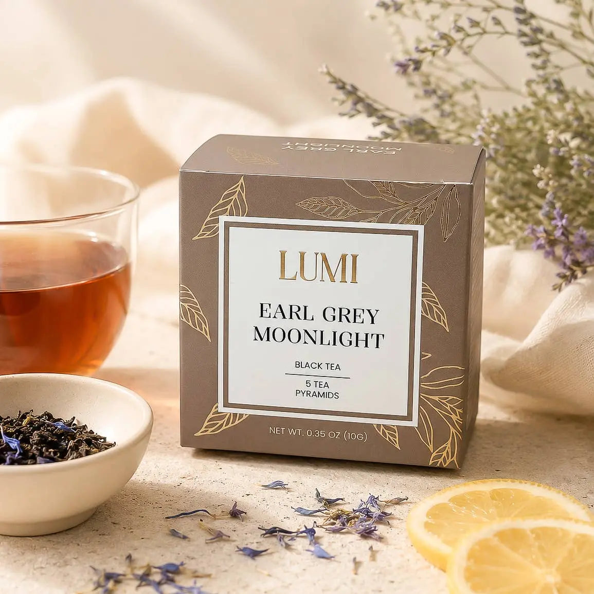 A box of LUMI Earl Grey Moonlight black tea sits on a light stone surface with a glass of brewed tea, a small bowl of loose tea leaves, dried flowers, and lemon slices around it.