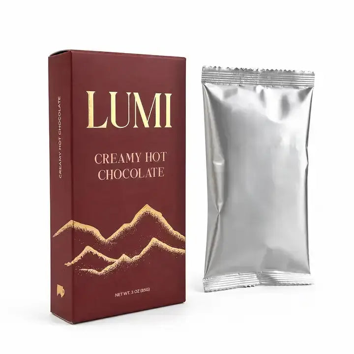 A box of Lumi Creamy Hot Chocolate next to a single silver foil packet on a clean white background.