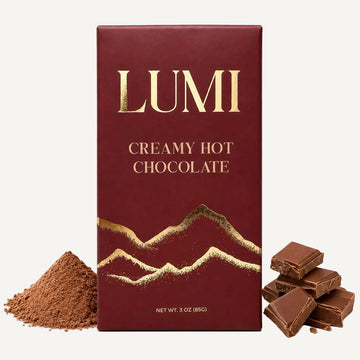 A 3 oz box of LUMI Creamy Hot Chocolate in its burgundy and gold wrapper, displayed with a pile of cocoa powder on the left and several broken chocolate pieces on the right, all on a clean white background.
