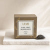 LUMI Earl Grey Moonlight black tea box in a warm brown tone with gold leaf illustrations, displayed on a white stone slab next to a single pyramid tea bag containing loose tea leaves.