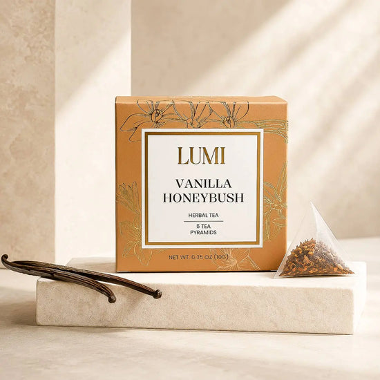LUMI Vanilla Honeybush herbal tea box in a warm tan color with gold floral accents, displayed on a light stone pedestal with vanilla beans and a tea pyramid packet.